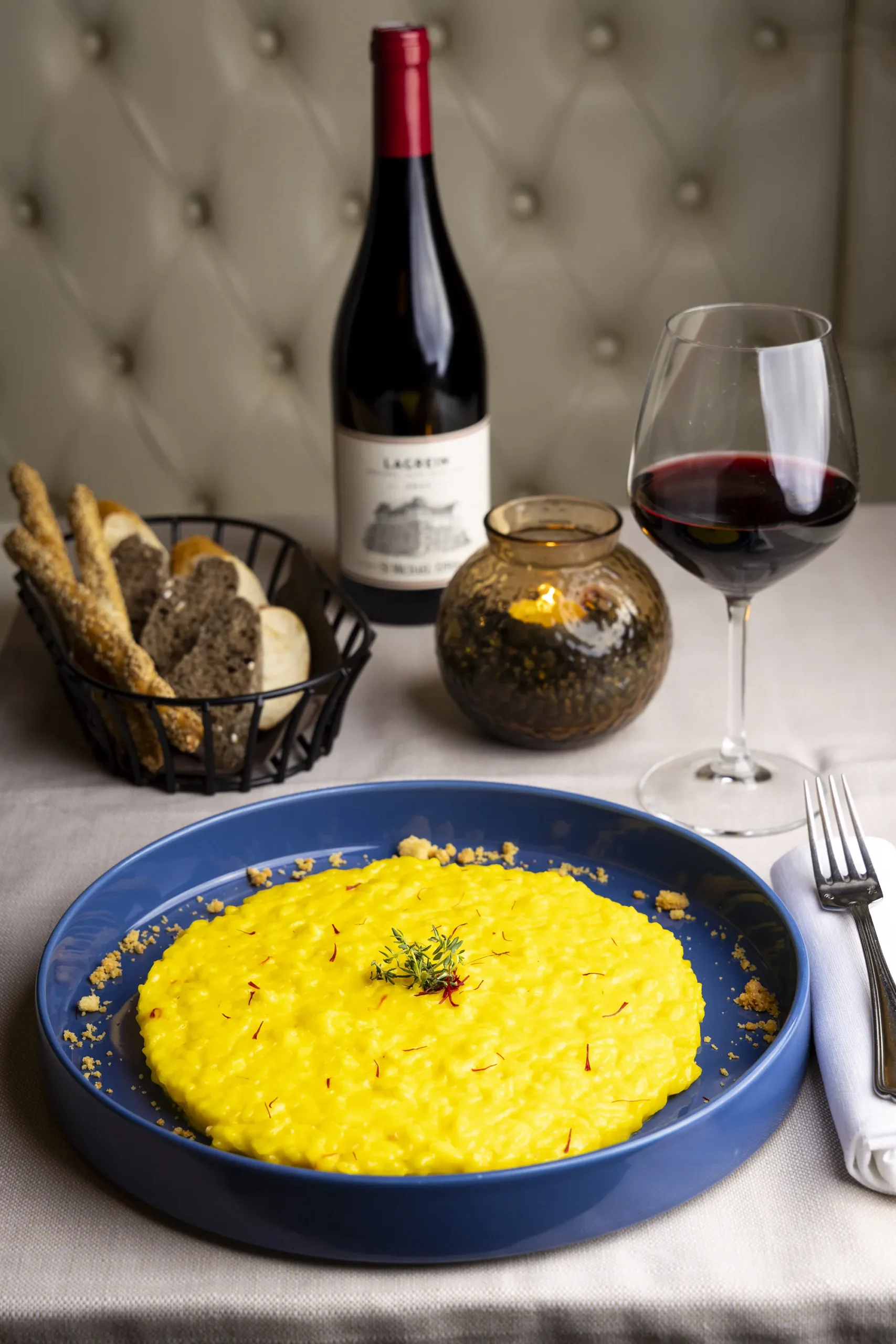 Un piatto di risotto allo zafferano, con un bicchiere di vino rosso e sullo sfondo una bottiglia di vino e un cestino di pane