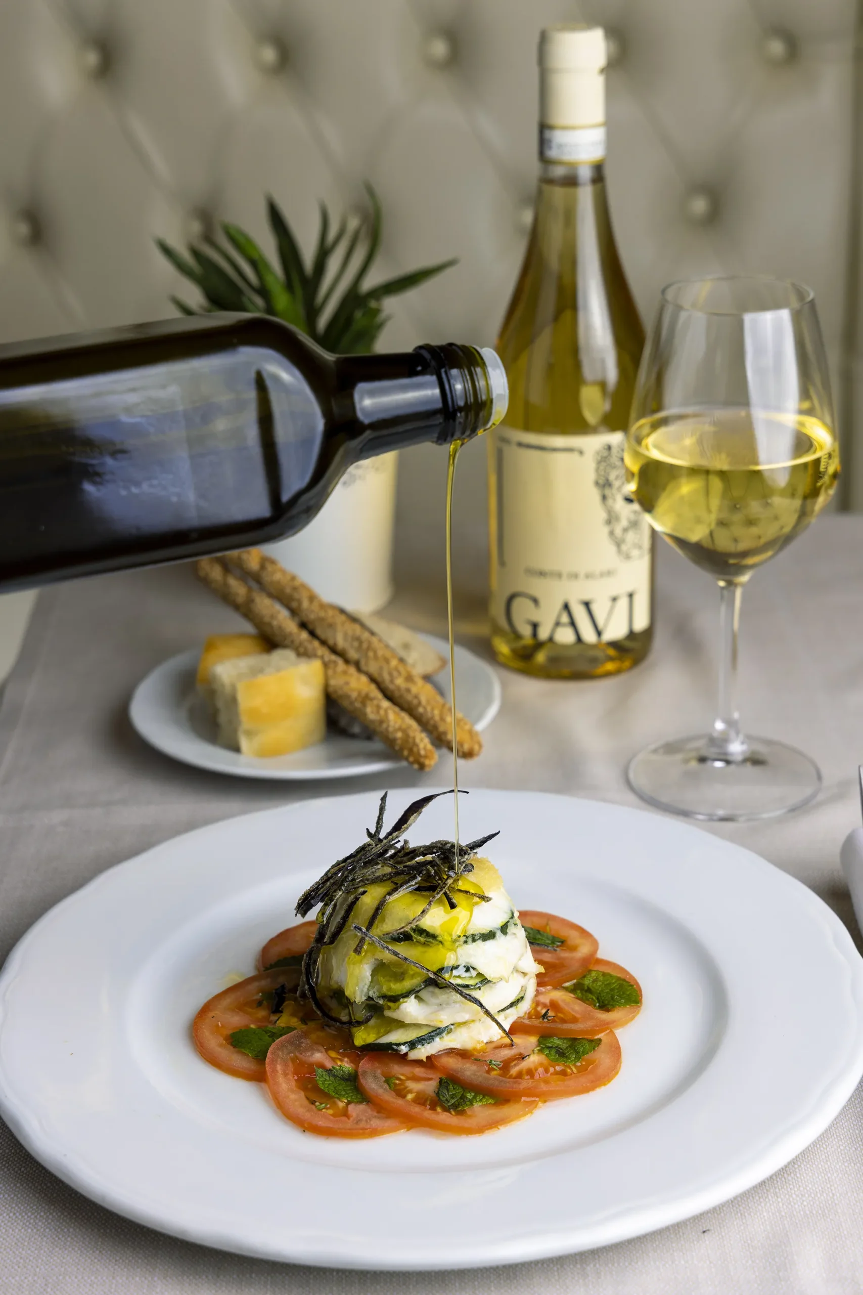 Un piatto con un tortino di verdure su letto di pomodori e, sullo sfondo, dei grissini, un bicchiere di vino bianco e una bottiglia di vino. Una mano versa dell'olio a filo sul piatto.