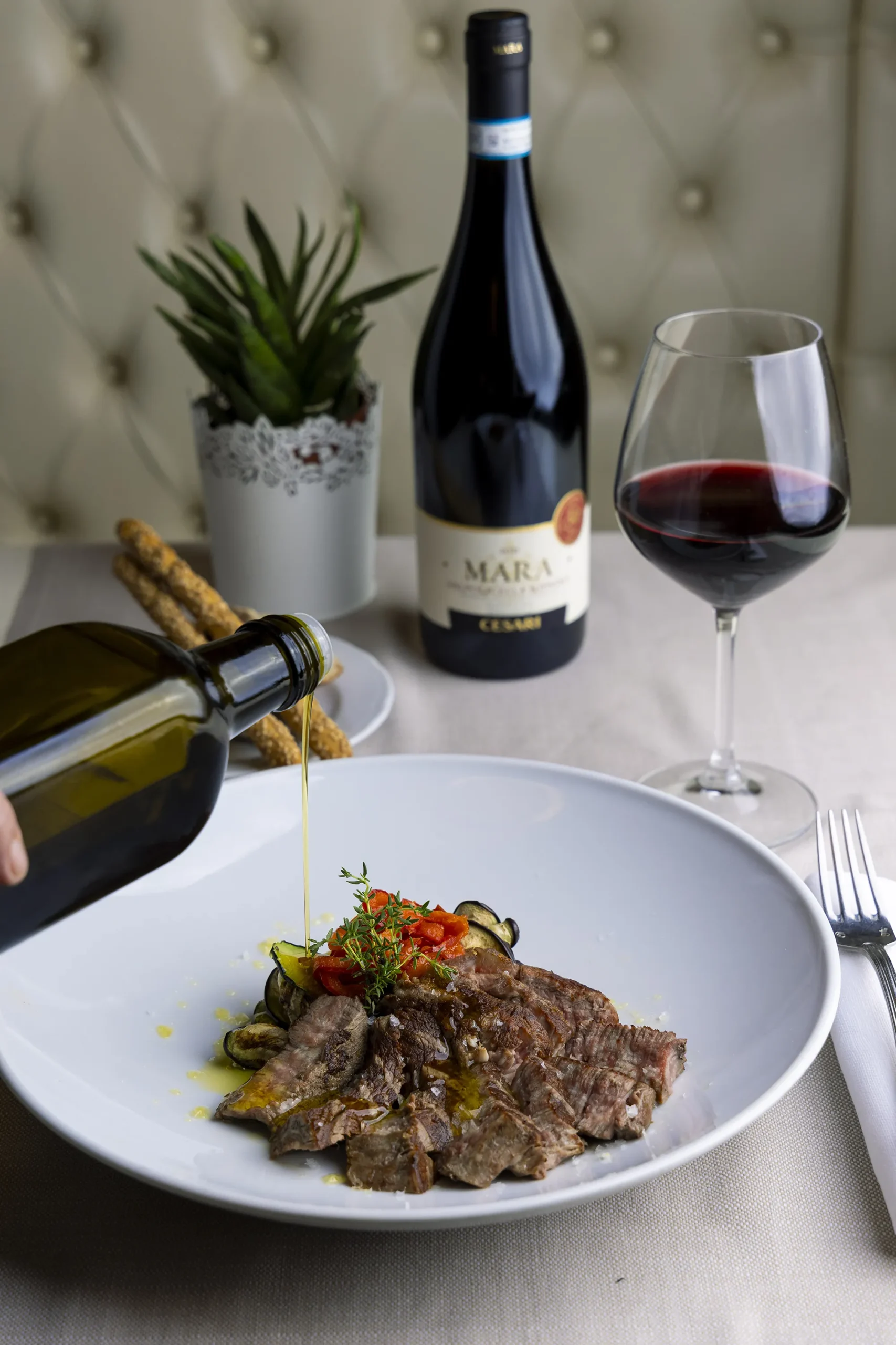 un piatto con delle fette di carne alla griglia, su cui viene versato dell'olio. Sullo sfondo una bottiglia di vino ed un bicchiere con del vino rosso