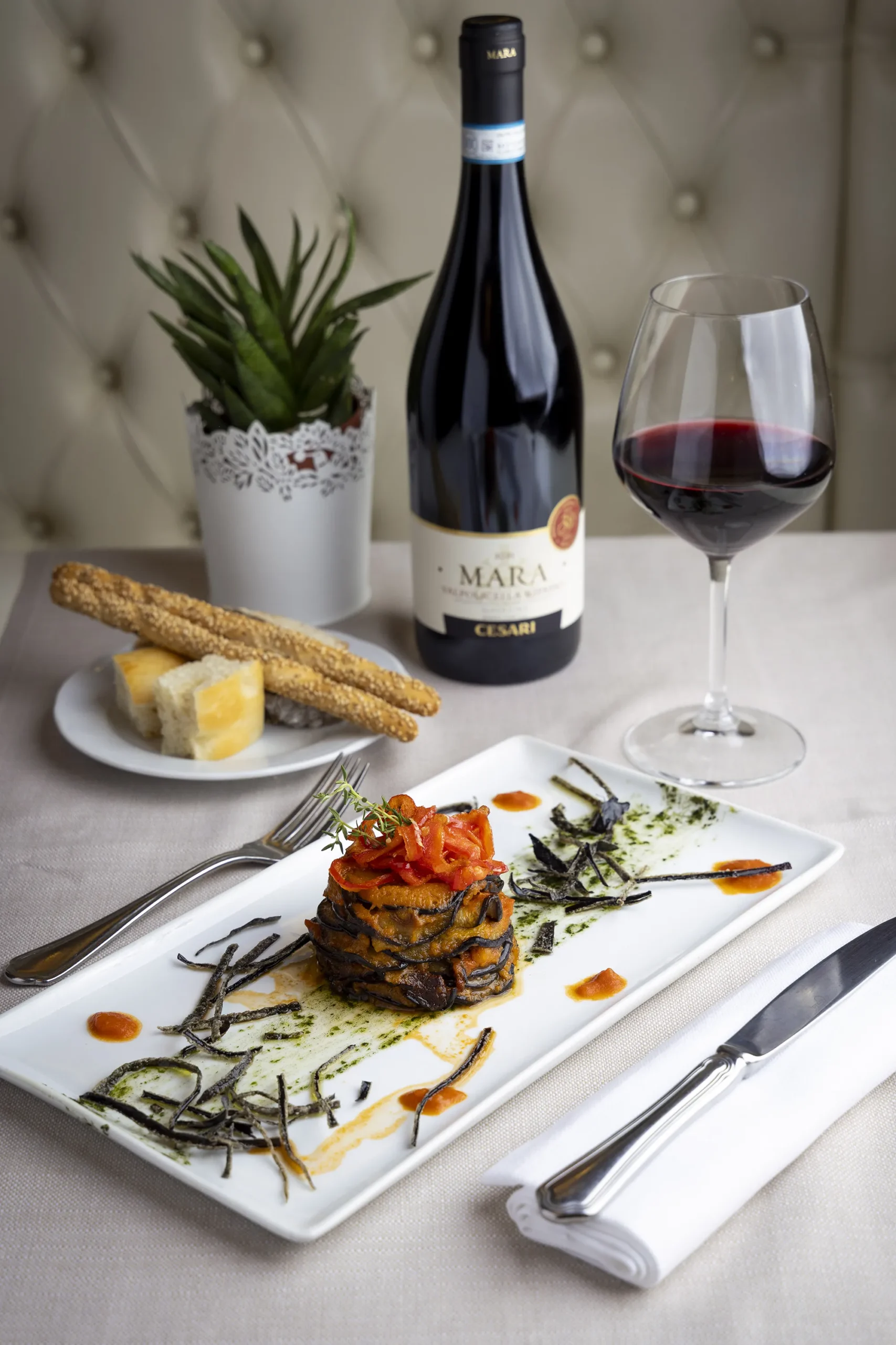 Un piatto con un tortino di melanzane, sullo sfondo un bicchiere di vino rosso, una bottiglia e un piatto con dei grissini